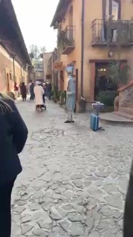 The video shows a paved street with a performer standing still in a blue and white costume, including a headpiece that resembles a glass container with a yellow object inside. The performer is wearing black gloves and formal shoes. Behind the performer, there are buildings that resemble European architecture with balconies and a sign that says "THE ARK". People are walking in the background. There is a suitcase and a small stand next to the performer.  The day appears to be bright and sunny. The video is shot from a slightly low angle.
