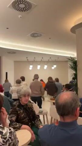 The video shows a group of older adults dancing together in a brightly lit room. Some are seated, watching the others dance. The room has a modern aesthetic with light-colored walls, modern lighting fixtures, and potted plants. The dancers are dressed casually and appear to be enjoying themselves.  Those seated are in small groups around tables, engaged in conversation.