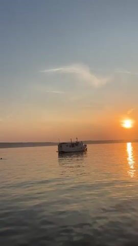A wide shot shows a boat on a calm body of water during sunset. The sun, a bright orange orb, is low on the horizon, casting a shimmering reflection on the water. The sky transitions from a clear blue at the top to a soft orange and pink near the horizon. A small, white boat with a cabin is positioned in the middle ground of the image. The overall scene depicts a peaceful, serene evening on the water.