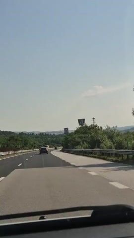 The video shows a view from a car driving on a highway. The road is asphalt with white lane markings. There are trees and vegetation on the right side of the road. In the distance, there are hills and a clear blue sky. There are a few other cars on the road ahead. On the right side of the road, there is a tall sign with the KFC logo. There is also another sign with a circular symbol. A utility pole with wires is visible behind the KFC sign. The camera is inside the car, and the windshield wipers are visible at the bottom of the frame.