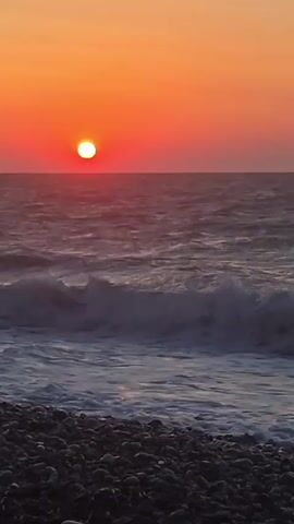 A vibrant sunset over a choppy ocean is captured in this vertical video. The sun, a bright orange orb, is positioned on the left side of the frame, casting a warm glow across the sky which transitions from a deep orange near the horizon to a lighter hue above. The sea below is a deep blue-gray, with white-capped waves rolling towards the shore. The foreground of the image is a dark, pebbly beach, with the waves lapping at the stones. The video appears to be a still shot or has very minimal movement, focusing on the natural beauty of the sunset and the ocean. There are no people or man-made objects visible in the scene.