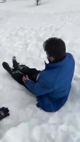 A group of people is playing in the snow in an open, hilly field with sparse trees. One man in a blue puffer jacket slides down a snow-covered hill on his back while others run in the background. One person walks toward the camera holding a large clear plastic bag. The snow covers the ground completely, and there are patches of bare earth visible. The weather appears overcast. The camera is mostly still but slightly shaky.