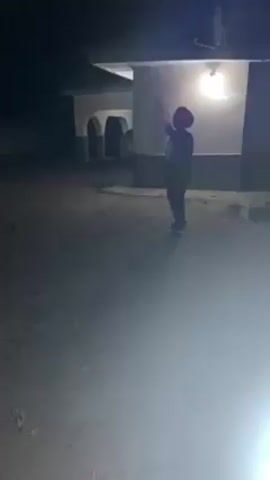 A person wearing a dark outfit and a red head covering is standing in a courtyard at night. The person is facing away from the camera and towards a building. The building has a light fixture illuminating the courtyard, and a set of arches is visible on the left. The ground appears to be a mixture of dirt and gravel. The camera is positioned low to the ground, and there is a bright light source in the foreground. The overall lighting is dim.