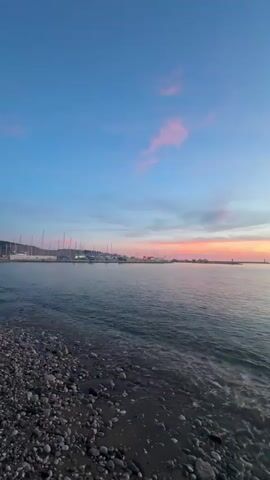 The video shows a calm sea at sunset. The sky is a mix of blue and pink. There are some clouds in the sky. The water is gently lapping on a rocky shore. In the distance, there are some boats docked in a harbor. The sun is setting behind some mountains, casting a warm glow over the scene.