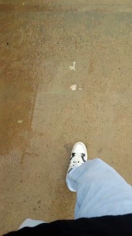 The video begins with a close-up of a person's foot wearing a white sneaker with black stripes and light blue jeans walking on a wet concrete sidewalk. The camera angle then shifts to a wider view, revealing the person walking along the sidewalk, which runs alongside a street wet from rain. A chain-link fence topped with barbed wire borders the sidewalk on the left, with green foliage growing behind the fence. Utility poles with overhead wires line the street. Buildings are visible in the distance, and a hill is seen in the background under a cloudy sky. The street appears empty except for the person walking.