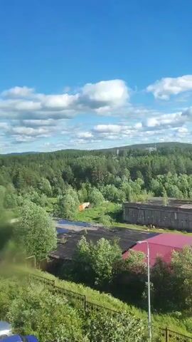This media file captures an outdoor scene with a clear blue sky dotted with white, fluffy clouds, indicating pleasant weather, likely during the daytime. The camera pans across a landscape that appears to be on the outskirts of a settlement.

In the foreground, a black metal fence runs horizontally, partially obscured by lush green foliage and bushes. Beyond the fence, a building with a dark roof is visible, adjacent to a structure with a prominent red, corrugated metal roof. A tall, white lamppost stands near the red-roofed building.

The midground reveals more industrial or utilitarian buildings. One long, gray, concrete structure with a dilapidated appearance is prominent, showing signs of wear and tear. A few trees are scattered around this area. Further back, more buildings, some with red roofs, are interspersed with dense green trees forming a forest that extends to the horizon.

In the background, a tall, dark smokestack rises from a building, suggesting an industrial facility. Beyond that, the landscape is dominated by a vast expanse of green forest, with rolling hills in the distance. Several other structures, possibly part of a town or industrial complex, can be seen amidst the trees and on the distant hills.

The overall impression is one of a somewhat undeveloped or industrial area blending with nature. The presence of factories or industrial buildings alongside residential-looking structures, all surrounded by extensive woodland, suggests a location that might be a town or village in a forested region. The clear sky and daytime lighting indicate favorable conditions for observation. There are no people visible in the frame. No discernible text is present. The location context provided, Alakurtti, Russia, aligns with the visual cues of a northern, forested environment with industrial elements.