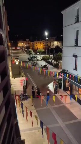 The video shows a street scene at night, decorated with colorful flags strung across it. There are several buildings lining the street, some with shops on the ground floor, their entrances illuminated. Pedestrians are visible walking along the street and on the sidewalks. A few people are standing and interacting in small groups. There are also parked motorcycles and bicycles. The lighting suggests it is evening or night, with streetlights and lights from the buildings illuminating the area. The overall impression is of a lively urban environment with some festive decorations.