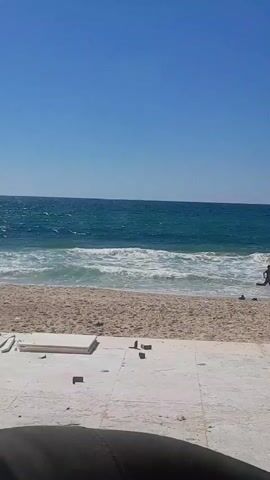 The video shows a view of a beach with the ocean in the background. The water is a deep blue-green and there are white waves breaking on the shore. The sand is light brown and there are some scattered objects on the sand, including what appear to be construction materials like bricks and possibly some white sheeting. The sky is a clear, bright blue. The lower portion of the video is obscured by a dark, blurry object, possibly a person's leg or clothing. There are no distinct actions or interactions visible beyond the natural movement of the waves.