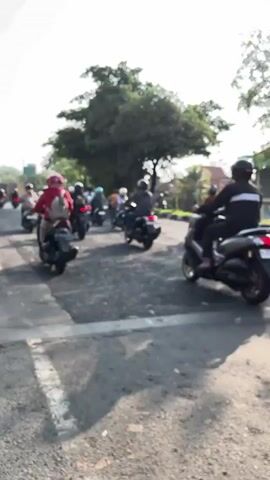 The video shows a street scene with several motorcycles traveling in the same direction. The motorcycles are on a paved road with gravel shoulders. There are trees and buildings visible in the background. The sky is clear and the sun is shining. The video is filmed from the perspective of someone riding on a motorcycle, looking ahead. The first shot shows a line of motorcycles, with the closest one on the right side of the frame. The second shot shows a motorcycle with two people on it, traveling in the same direction. The third shot shows the same motorcycle from a slightly different angle, with the person in the back wearing a backpack. The video appears to be filmed during the day, likely in the morning or afternoon.