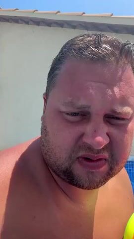 A close-up video shows a man in a swimming pool holding a colorful inflatable ball. He is partially submerged in the blue water, with his shoulders and chest visible. The man has dark hair and a beard, and his skin appears flushed, possibly from the sun. The background consists of the pool's tiled edge and a light-colored wall. The man looks directly at the camera and smiles, then his expression changes to a frown, before returning to a smile. The inflatable ball, with its red, orange, yellow, and green sections, is held on his right side. Sunlight illuminates the scene, creating shadows and highlights on the water and the man's skin. The man appears to be enjoying himself in the pool, as indicated by his smiles and the presence of the beach ball.