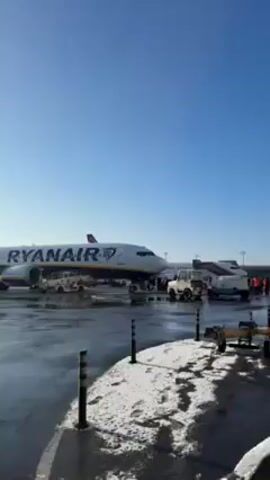 The video shows an airport scene on a sunny day with patches of snow on the ground. A Ryanair plane is visible on the tarmac. Passengers are disembarking the plane. The camera pans towards a modern building with large windows, likely an airport terminal. A line of people is waiting to enter the building. A man wearing a brown jacket and a black cap walks towards the line. The camera follows him as he approaches the entrance.