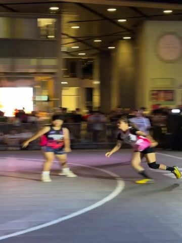 The video shows a women's basketball game being played outdoors at night. The players are wearing pink and black jerseys. The court is surrounded by a metal fence and a crowd of people. In the background, there is a building with the sign "Ladurée" and "National" visible. One player attempts a shot and scores.