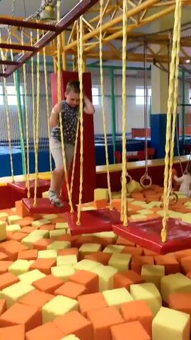 This video shows a young boy navigating a rope course in an indoor playground. The boy, wearing a t-shirt and shorts, is on a suspended platform, gripping a rope for balance as he moves across the course. The platforms are red and suspended by yellow ropes from a metal structure overhead. Below the course is a large pit filled with orange and yellow foam cubes, designed to cushion falls. In the background, other playground equipment is visible, including what appears to be a blue climbing wall or net, and gymnastic rings. The setting appears to be well-lit, suggesting it is daytime indoors. The boy's expression is focused as he concentrates on his movement. The overall scene depicts a recreational activity in an amusement facility.