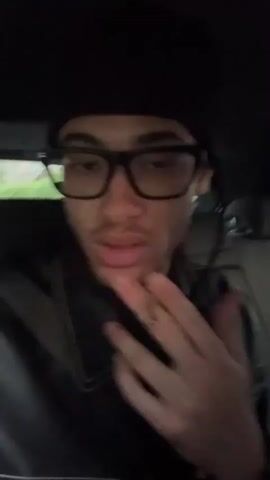 A young man with dark hair in braids, wearing a black beanie, black-framed glasses, and a black leather jacket with white stitching, is sitting in the driver's seat of a car, smoking a cigarette. The car interior is grey. He looks directly at the camera. Outside the car, a blurry image of a house is visible.