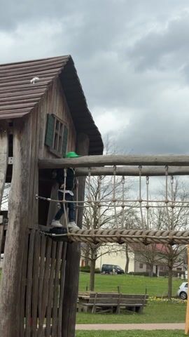 A young child wearing a black jacket, jeans, white sneakers, and a green baseball cap walks across a rope bridge connecting two wooden structures in a playground. The structure on the left is a small, rustic wooden house with a brown shingled roof and a small green shuttered window. The rope bridge is made of thick rope and metal cables. The playground is located in a park-like setting with green grass, trees, and buildings visible in the background. The sky is overcast and gray.
