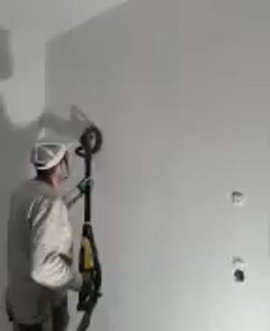 A man is sanding a white wall with a long-handled, circular sander. He is wearing a white painter’s hat, safety glasses, a long-sleeved grey shirt, and gloves.  The room has a corner, and the wall has two electrical outlet boxes without covers.  The shadow of the man and sander is visible on the wall to the left.