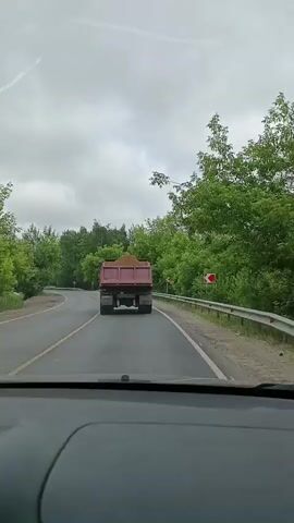 A first-person perspective video shows a view from inside a car looking forward at a red dump truck driving on a paved road. The dump truck is carrying a load of dirt or sand. The road curves to the left ahead, and there are green trees and foliage on both sides of the road. There are guardrails along the right side of the road, and a red and white traffic sign indicating a curve is visible. The sky is overcast.