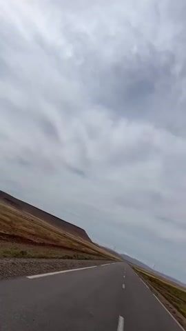 The video depicts a view from a moving vehicle traveling on an asphalt road through a rural landscape on a cloudy day. The road is straight and stretches into the distance, marked with a white lane divider. On either side of the road, the terrain is primarily brown and dry with sparse vegetation. A long, low plateau runs along the horizon on the left side of the frame. In the far distance, what appear to be utility poles and faint mountains are visible. The sky is overcast with thick gray clouds.