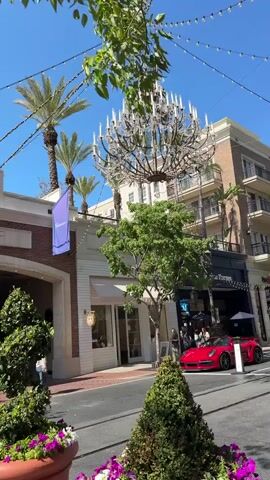 The video captures an outdoor street scene with a large chandelier hanging overhead. Palm trees and other greenery are visible, along with buildings that appear to be shops or restaurants. A red sports car is parked on the street. Several people are walking along the sidewalk. The sky is clear and blue, suggesting a sunny day.