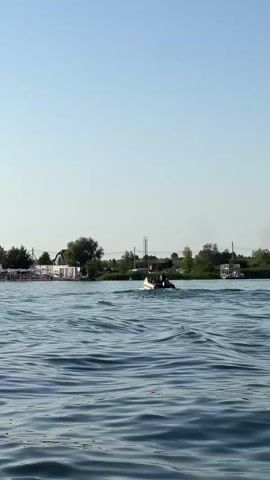 A wide shot captures a serene daytime scene on a body of water. The water is a calm blue, with gentle ripples disturbing its surface. In the middle ground, a small white boat with a few people on board navigates the water, creating a small wake.

Along the distant shoreline, a line of white umbrellas and structures suggests a beach or recreational area. Behind this, a scattering of trees and buildings are visible under a clear, pale blue sky. The overall atmosphere is peaceful and summery, suggesting a relaxed day outdoors. The time of day appears to be daytime, likely late morning or early afternoon, given the bright, even lighting. The location is identified as Verbizh, Ukraine. There is no visible text in the image.