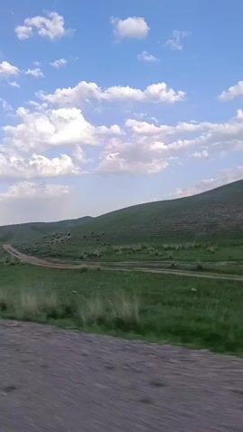 The video shows a dirt road winding through a grassy, hilly landscape under a blue sky with scattered white clouds. The camera is moving, creating a sense of motion. There are some dark shapes on the distant hills that could be animals or rocks. A white road marker is visible on the right side of the road, followed by a concrete barrier. The sun appears to be low in the sky, casting a warm light on the scene.