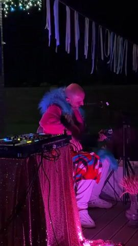 A bald person wearing a pink hoodie with blue fur trim and light-colored pants sits on a wooden deck at night, facing a microphone. To the left, a DJ setup with flashing lights sits on a table draped in a pink sequined cloth. A person with dark hair is partially visible behind the main subject, holding a phone. Decorative ribbons hang from an overhead structure. The environment appears to be outdoors with some greenery in the background.
