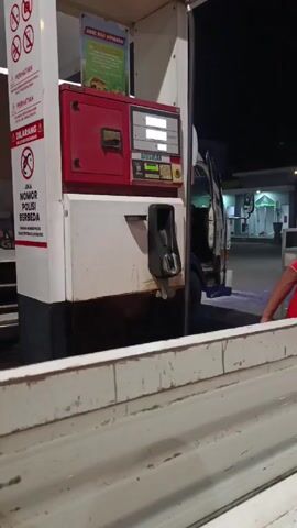 The video depicts an outdoor gas station at night. A white pickup truck with an open passenger door is parked next to a gas pump labeled "BIOSOLAR." A man in a light green shirt and dark shorts is walking around the truck near the gas pump. The foreground shows part of the side of another white vehicle, potentially another pickup truck. The lighting indicates it is nighttime with overhead lights illuminating the gas station. A building with a glass facade is visible in the background.