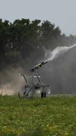 A large irrigation sprinkler is positioned in a field of green crops. The sprinkler is actively spraying a powerful stream of water into the air. Behind the sprinkler, a green tractor is visible, partially obscured by a cloud of dust or mist generated by the irrigation process. A dense line of dark green trees forms the background. The sky is overcast, suggesting cloudy or hazy weather conditions. The overall scene depicts agricultural irrigation in progress.