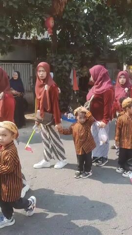 A group of children and adults are participating in a parade. The children are dressed in traditional striped shirts and pants, with some wearing head coverings. They are holding small flags and walking in a line on a paved road. The adults, mostly women, are wearing red headscarves and matching outfits. They appear to be supervising the children. The background shows trees, houses, and a fence. The overall atmosphere seems festive and celebratory.