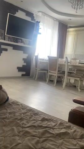 The video shows a view of a room, possibly a living or dining area. The camera is positioned low, looking up towards a dining table and chairs set near a window. The window has sheer white curtains and darker drapes. To the left, a large flat-screen television is mounted on a wall decorated with a textured, brick-like pattern. The floor is light-colored laminate. In the background, a white kitchen cabinet area is visible. The foreground shows a portion of a bed or sofa with a quilted, beige throw blanket. The lighting suggests it is daytime, with bright light coming from the window.
