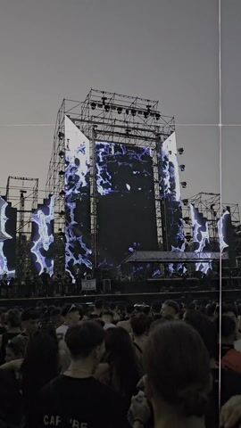The video shows a large outdoor stage with a crowd of people in front of it. The stage is elaborate, with large video screens displaying a swirling blue and white pattern. The crowd is mostly young adults, many wearing dark clothing. The lighting is dark and atmospheric, suggesting it is evening or night. The overall mood is energetic and festive. The stage setup indicates a music concert or festival.