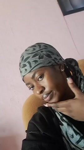 A young woman with dark skin and braids, wearing a gray and black headscarf, a black top, and small butterfly earrings, sits in an upholstered tan chair. She looks directly at the camera, touching her chin, briefly pursing her lips, and then adjusting the scarf around her neck. The background includes a pale pink wall and a framed picture hanging slightly out of view.