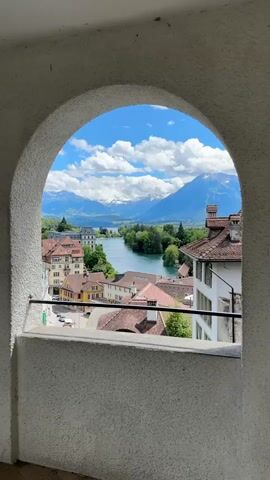 The video shows a view through an arched window of a town situated along a river, with mountains in the background. The town has buildings with red-tiled roofs and stucco walls, some with balconies. A river flows through the middle of the town, bordered by trees. In the distance, snow-capped mountains rise against a blue sky with white clouds. The camera appears to be stationary, and there are no people or discernible actions occurring in the scene. The lighting suggests it is daytime, likely a clear and sunny day. The overall impression is a scenic, picturesque landscape.