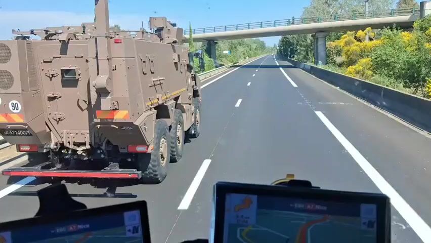 The video shows a military vehicle traveling on a highway. The vehicle is a brown, boxy, armored truck with a crane on top. It has the numbers "80" and "90" displayed on the back, along with a license plate number. The vehicle is driving in the right lane of a highway with white lines. The view is from behind, as if the camera is in another vehicle following the military truck. The highway is surrounded by trees and vegetation. There are a few overpasses visible in the distance. The sky is blue and the sun is shining. Two GPS devices are visible in the foreground, mounted on the dashboard of the camera vehicle. The road looks to be in good condition.