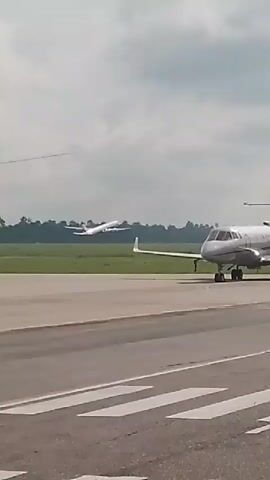 The video shows an airport tarmac under a cloudy sky. In the foreground, a silver private jet is parked on the tarmac. In the background, another airplane is in the process of taking off, ascending into the sky. The tarmac has clear white markings. The scene is captured from a stationary perspective, possibly from another vehicle or a fixed point on the ground.