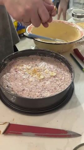 A close-up, top-down shot shows a person's hands crushing small pieces of what appears to be cookie or biscuit into a round, dark baking pan. The pan is filled with a pinkish, creamy mixture containing small red flecks, possibly berries. The person's fingers are visible, breaking off pieces of the topping and sprinkling them over the surface of the mixture. In the background, a yellow bowl with a white rim is visible, as well as a red-handled knife lying on a white surface. Other blurred figures and objects are also present in the background, suggesting a kitchen environment. The lighting is bright, illuminating the actions on the cake.
