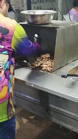 A man wearing a colorful cartoon shirt and black gloves is cooking. He is inside of a mobile food stand. He is using a spatula to flip grilled meat inside of a stainless steel hood. To the right of the grill are two deep fryers, one contains french fries and the other contains hotdogs. A blue bowl with tomatoes sits behind the fryers, along with a container of condiments. There is music playing in the background.