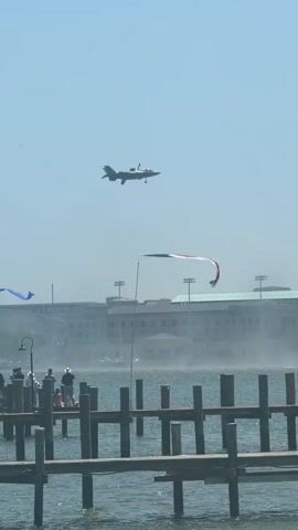 A gray fighter jet approaches to land, flying in the clear blue sky over a body of water and a wooden dock.  The jet has its landing gear down and is angled slightly down, appearing to be on its final approach. The dock has people standing along its edge, facing the plane. Beyond the dock is a building with multiple levels and many windows. Three flags, one blue, one American and one red, are mounted on poles that appear to be on the dock. The surface of the water is slightly choppy, and the sun is bright.