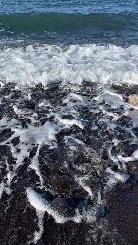 The video shows a close-up view of waves crashing on a dark, rocky beach. The water is foamy and white as it washes over the black sand and stones. The camera is positioned low to the ground, giving the viewer a perspective close to the water's edge. The scene is natural and peaceful, with the sound of the waves creating a calming atmosphere. The rocks on the beach vary in size and shape, and some are smooth and rounded, while others are rough and jagged. The water appears to be clear, with a slight greenish tint.