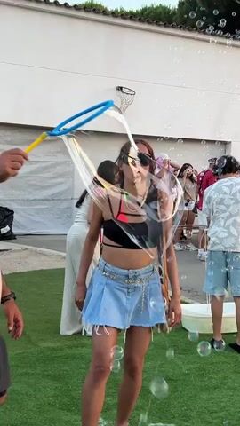 A young woman with dark hair and sunglasses, wearing a black crop top, denim skirt, and sandals, is blowing large iridescent soap bubbles with a wand. She is in a grassy outdoor area with a group of people in the background, some of whom are watching her. The background also shows a building, a basketball hoop, and a white tub. The woman is focused on blowing the bubbles, and several are floating around her. The scene appears to be a casual gathering or event.
