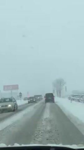 The view from inside a vehicle driving on a snow-covered road shows several cars driving ahead in the same direction. Snow is actively falling and visibility is poor. The road is mostly clear but has snow on the edges and packed snow in the tire tracks. There is a billboard on the left side of the road and a tree on the right side. The overall atmosphere suggests difficult driving conditions due to the winter weather.