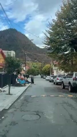 The video shows a quiet street in a small town. On the left side of the street, two men are standing near some construction materials. On the right side of the street, several cars are parked. In the background, there is a mountain covered in trees. The sky is partly cloudy. The street has several speed bumps.