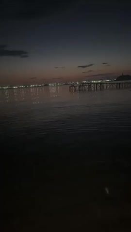 The video shows a body of water at dusk. The sky is dark with hints of pink and orange near the horizon. Lights from a distant city or town are visible across the water, reflecting on its surface. In the foreground, the water is dark and appears to have small ripples. A wooden pier or structure extends into the water on the right side of the frame. The overall atmosphere is calm and peaceful.