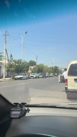 The video shows a street scene, likely from the perspective of someone inside a vehicle. The timecode indicates a duration of approximately 4 seconds. In the foreground, the dashboard of a vehicle is visible, with a black device mounted on it. The main focus of the video is the street and the vehicles on it.

On the right side of the road, a cream-colored van is parked. A person, appearing to be a male wearing a white t-shirt and light-colored pants, is standing by the open rear door of this van, facing away from the camera and looking down. Another similar cream-colored van is visible further down the road, also parked.

A line of other vehicles is present in the road, suggesting a traffic queue or parking. These include several white cars and what appear to be more vans. On the left side of the road, a white building and trees are visible. Utility poles with wires are also present. The sky is clear and blue, indicating a sunny day. The overall environment appears to be an urban or suburban street. The vehicles appear to be stationary or moving slowly.