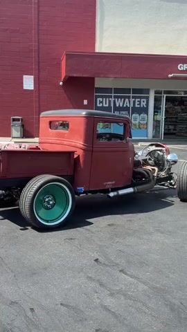 A vintage red hot rod truck is parked in a parking lot in front of a building with a red brick facade and a white upper section. The truck has a exposed engine, custom wheels with green rims, and a visible exhaust pipe. The building has a sign that says "CUTWATER" with pictures of canned beverages. A person is visible inside the driver's seat of the truck, and the truck appears to be stationary or moving very slowly. The ground is asphalt, and the lighting suggests it is daytime with bright sunlight.
