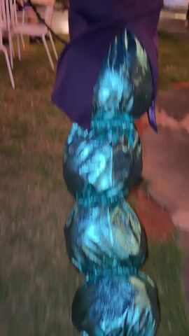 The video features a man in an elaborate costume at an outdoor evening event. He wears blue-grey face paint with darkened lines around his eyes, round green-tinted sunglasses, and a small patterned blue-green hat on his head. His attire consists of a long purple jacket, a white collared shirt, and a green necktie. A large silver watch is visible on his left wrist.

The man is holding a multi-colored, iridescent hookah, featuring shades of purple, blue, and green on its base, topped with a golden bowl and a black hose. He initially walks away from the camera, then turns to face it, and eventually places the hookah hose to his mouth, maintaining a slight, theatrical smirk.

The setting appears to be a garden or lawn area, with dark green grass underfoot. String lights with warm yellow bulbs are strung between trees, providing illumination against the dark blue dusk sky. Palm trees and other leafy trees are visible in the background. Several other people, mostly out of focus, are present at the gathering, some seated on white plastic chairs. The overall ambiance suggests a themed party or social event, consistent with a festive occasion in an outdoor setting like Nir Eliyahu, Israel. There is no visible text.