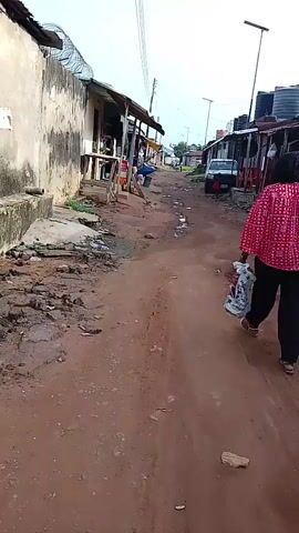 The video shows a woman walking away from the camera along a reddish-brown dirt road in what appears to be a market or residential area. She is wearing a red patterned top and black trousers, carrying several shopping bags in both hands.

On both sides of the road are modest, single-story buildings with tin roofs, many appearing to be small shops or stalls. Some buildings have faded, light-colored walls, and one has razor wire along its top. The ground is uneven, with patches of grass, small puddles, and scattered debris including plastic bottles.

In the background, a white vehicle is parked, and large black water tanks are visible on the roofs of some structures. Power lines stretch across the sky. Other people can be seen in the distance, including one person sitting outside a shop on the left side of the road. The sky is bright and clear, indicating daytime. The overall scene depicts a bustling, semi-urban environment in Ohokobe Ndume, Nigeria. No specific text is clearly legible on the shopping bags or buildings.