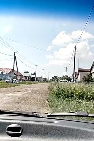 A view from inside a vehicle looking out at a dirt road in a rural village. There are several houses and wooden fences lining the road, with power lines overhead. The weather appears to be sunny with scattered clouds. The video appears to be a first-person perspective of someone driving through the village.