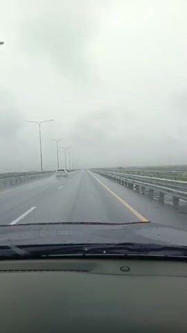 A POV shot from inside a car traveling on a wet highway in overcast conditions. The road has multiple lanes with white and yellow markings. Streetlights are visible along the side of the road, stretching into the distance. A white car is ahead, driving in the same direction. The windshield of the car has water droplets on it. The sky is completely obscured by clouds.