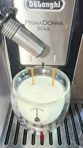 This is a close-up, medium shot video of an espresso machine pouring two streams of dark brown liquid into a clear glass mug. The mug is shaped like a polar bear's head, with the face printed on the outside. The liquid is being dispensed from an espresso machine with the brand name "DeLonghi" and model name "PRIMADONNA SOUL" visible on the front. The liquid is creating ripples in the white foamed milk that fills the mug. The shot is stable and focused on the mug and the espresso spouts.